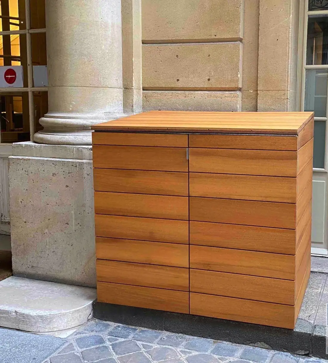 Coffre à conteneurs en lames d’iroko posé sous un porche d’immeuble