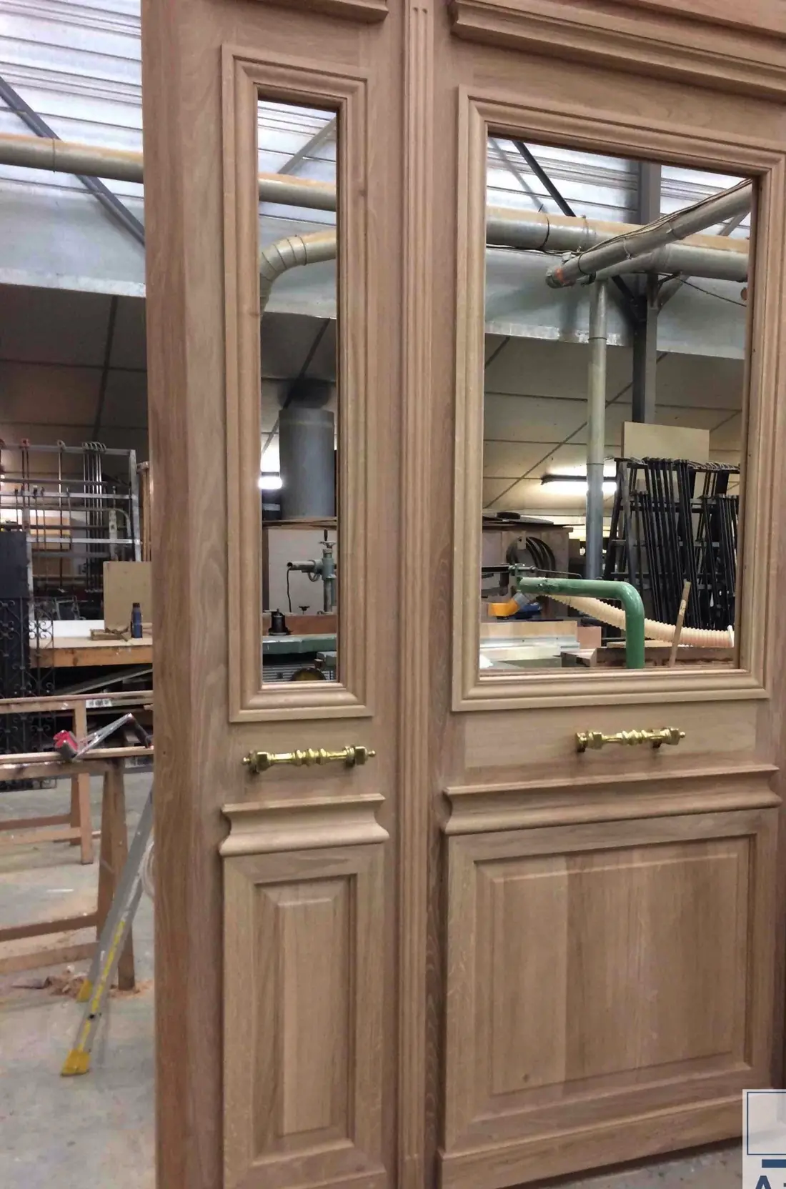 Porte à deux vantaux en chêne nu, vitrée en partie haute, debout dans l’atelier de Massy avant finition