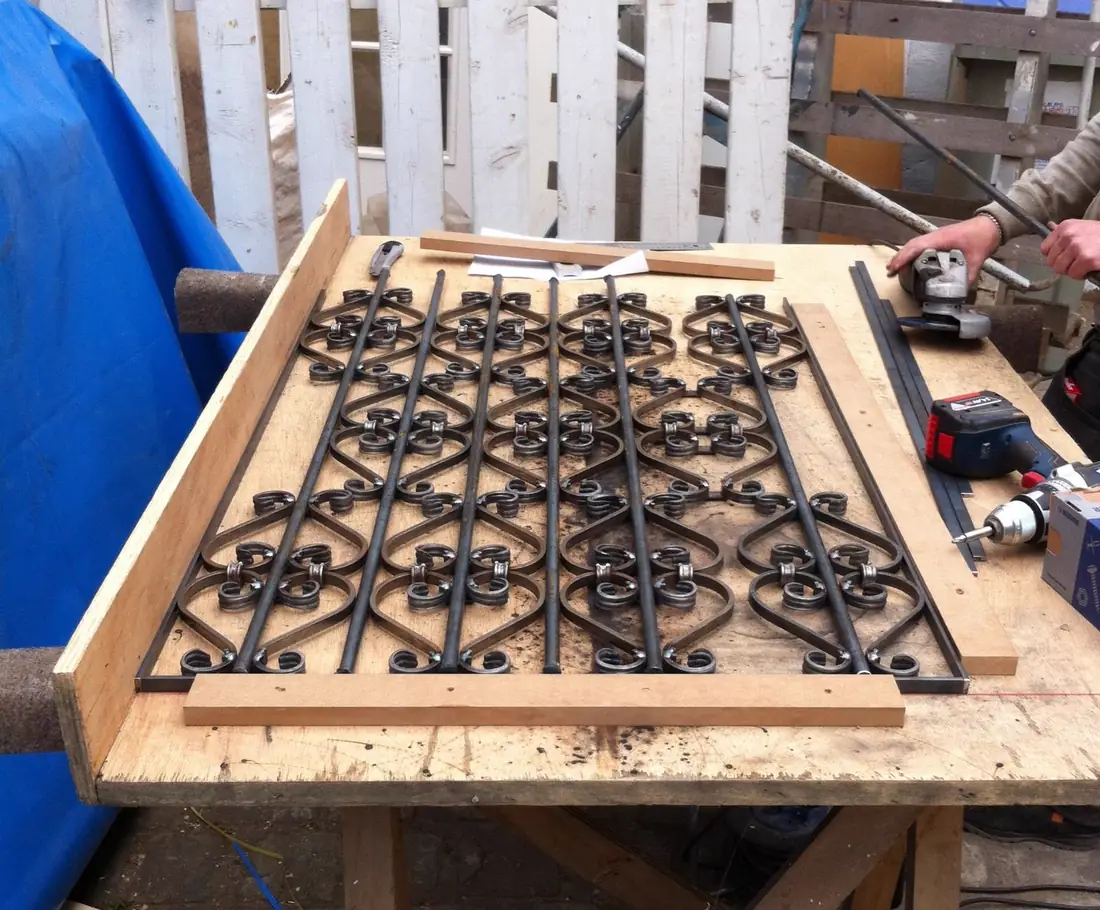 Grille à volutes serrée sur un établi, perceuse en main, pendant son façonnage