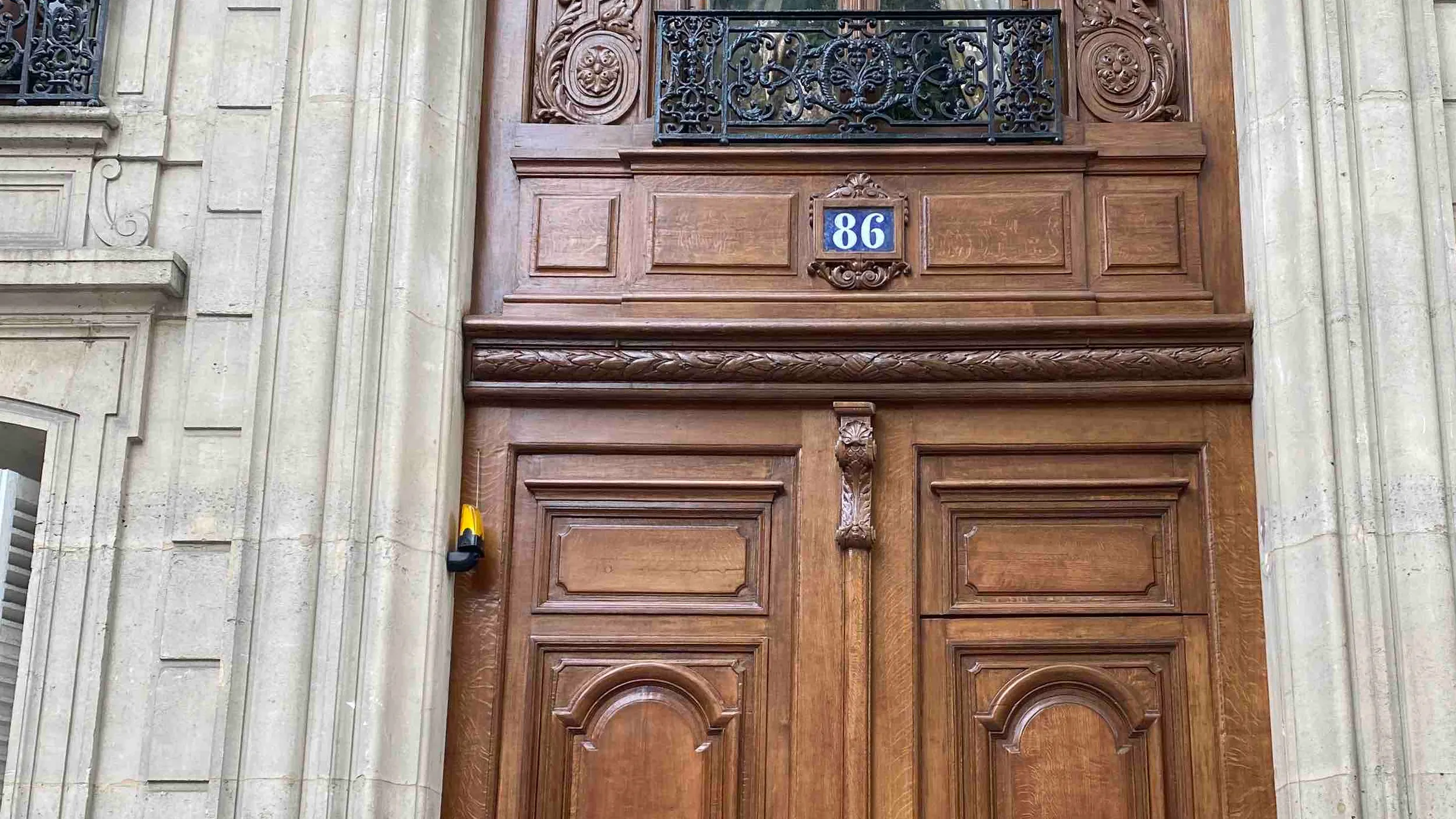 Porte cochère en chêne à deux vantaux, panneaux moulurés et consoles sculptées, sous une imposte vitrée à balcon en fer forgé et le numéro 86, dans une façade en pierre de taille
