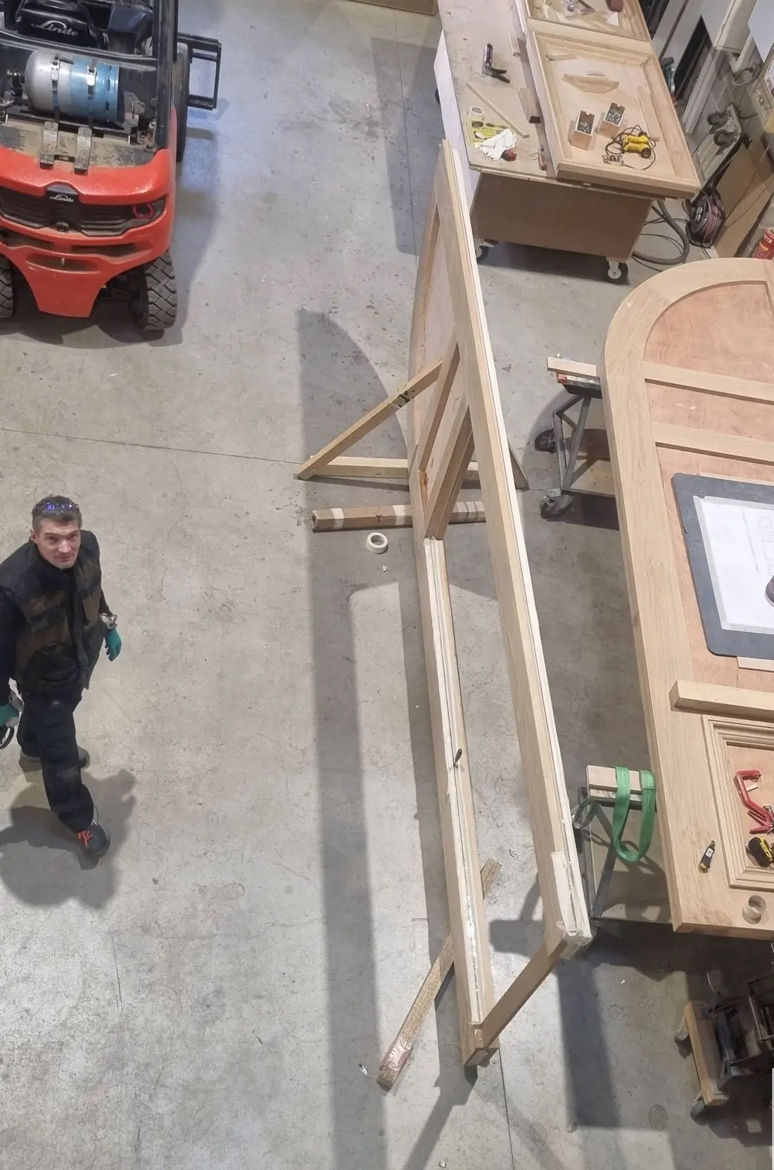 Vue plongeante de l’atelier : vantaux de chêne sur tréteaux, épure agrafée sur le panneau, compagnon au sol