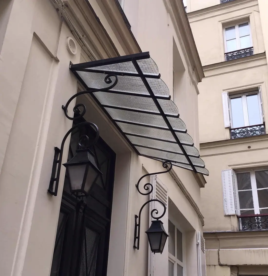 Marquise en acier et verre au-dessus d’une porte d’immeuble, encadrée de deux lanternes