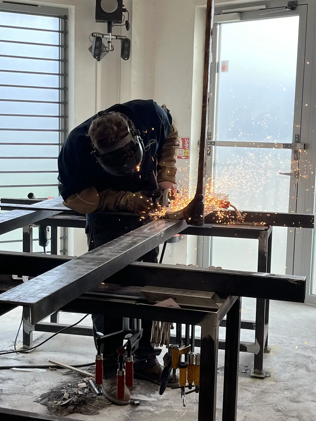 Compagnon meulant un profilé d’acier sur un établi de l’atelier, gerbe d’étincelles