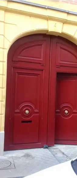 Porte cochère rouge cintrée à deux vantaux, portillon et boutons de laiton, sur une façade ocre