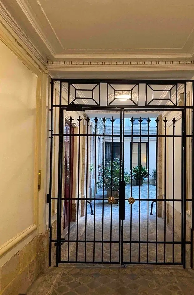 Grille de sas noire à barreaux et imposte à losanges fermant un porche, cour pavée au fond